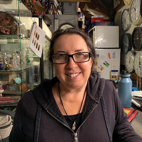 Photo of Samantha Gordon, Samantha looks smiling at camera and in background are a variety of props like clocks and trinkets